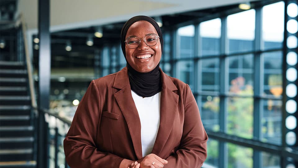 Mubeenat is standing and smiling and in focus. She is wearing a brown blazer and white t-shirt. In the background are big windows which are blurred.